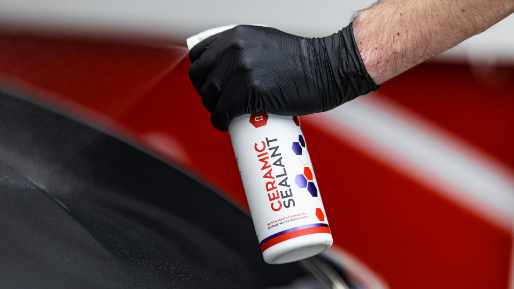 A person wearing black gloves sprays car polish onto a vehicle's surface.