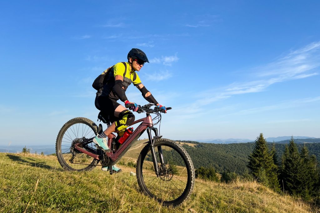 Cyclist man riding electric mountain bike outdoors. Male tourist biking along grassy trail in the mountains, wearing helmet and backpack. Concept of sport, active leisure and nature.